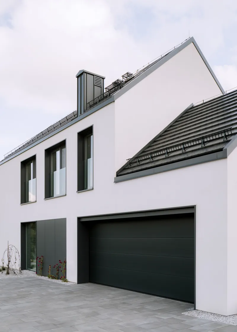 Street facade showing garage door and upper windows beneath intersecting gabled roofs with horizontal tile coursing