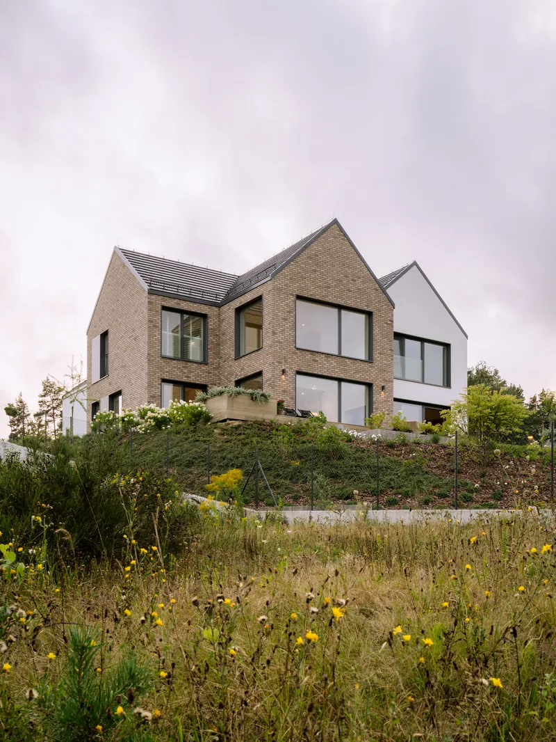Cluster of gabled brick and white-rendered volumes ascending a hillside meadow with yellow blooms