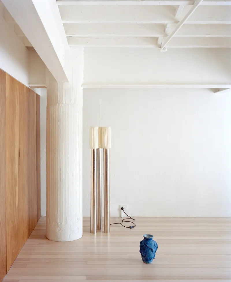 Open room with timber column, clustered copper pendant fixtures, and blue ceramic vessel on light wood flooring