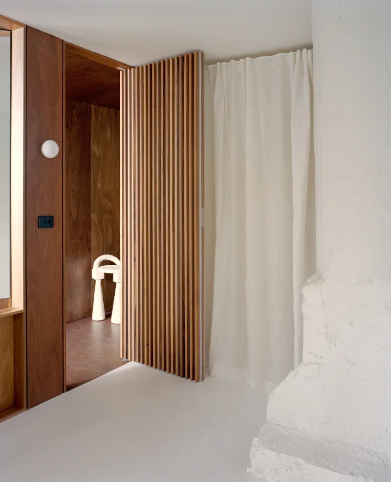 Bedroom threshold with vertical timber slat screen and white curtain dividing wood-paneled corridor