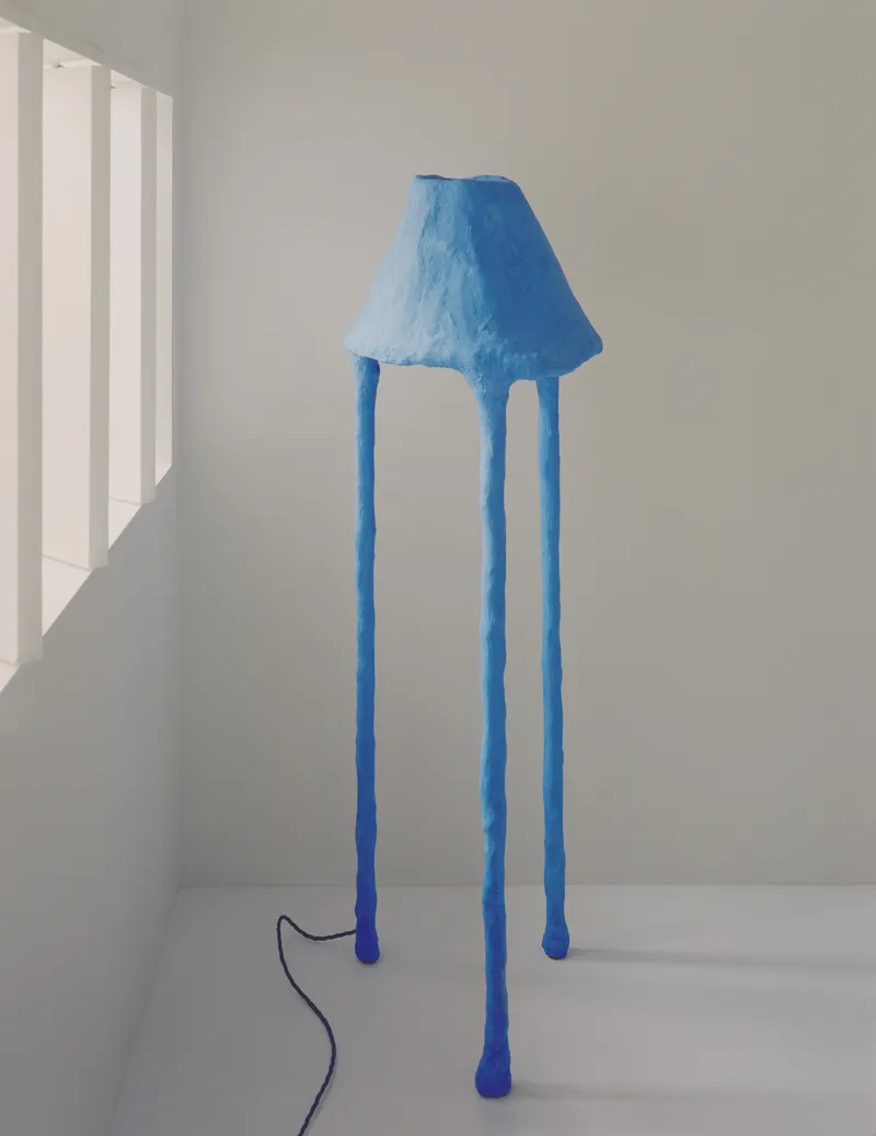 Blue tripod floor lamp with textured shade standing in a white corner with morning light