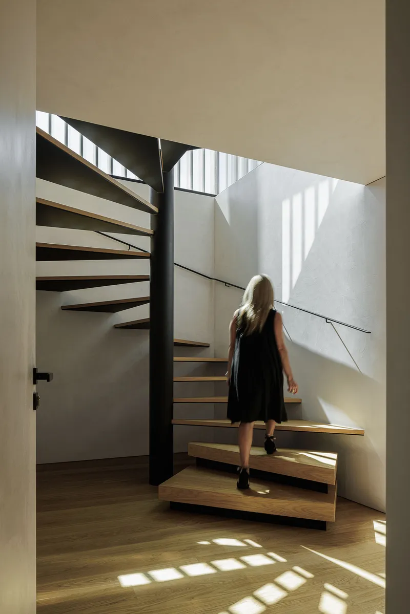 Timber and steel staircase with person ascending beneath a skylight casting geometric shadows across white walls