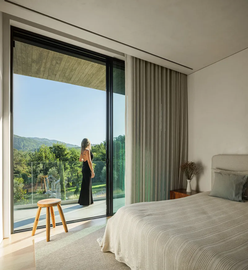 Bedroom with person standing at sliding glass doors overlooking the forested hillside in morning light