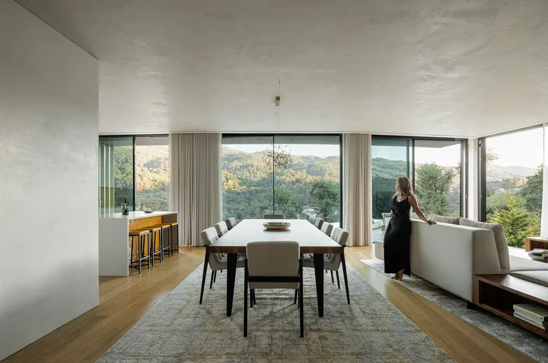 Open-plan dining area with continuous glass walls framing forested hills and a person at window