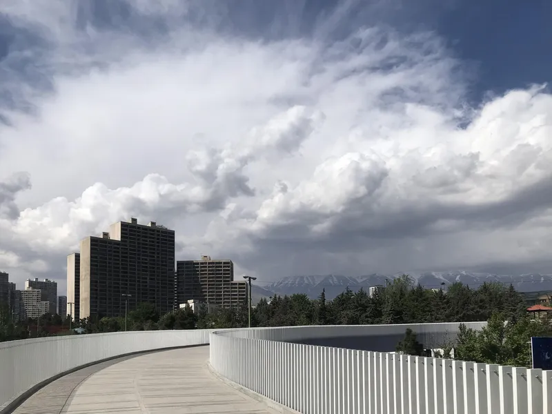 Curved pedestrian bridge with vertical metal siding overlooking residential towers and snow-capped mountains under storm clouds