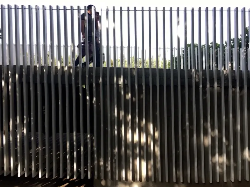 Figure walking behind vertical metal slats creating alternating bands of light and shadow