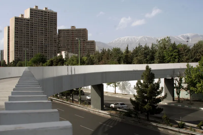 Open-tread staircase ascending alongside a pedestrian bridge with residential towers and snowy peaks beyond