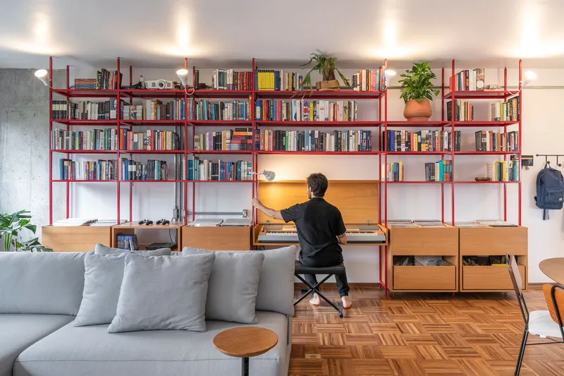 Red steel bookshelf wall with integrated desk where a person sits working at midday