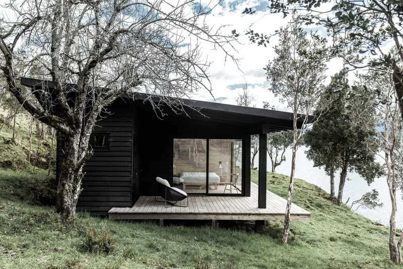 Black timber cabin with glazed facade and timber deck nestled on a grassy hillside among bare trees