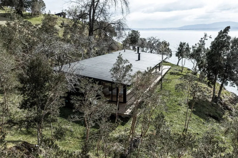 Aerial view of a low-pitched roof structure nestled among trees on a sloping hillside overlooking water