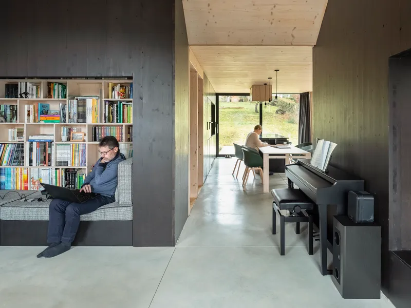 Interior view through concrete walls showing a man reading by the bookshelf and a woman at the desk