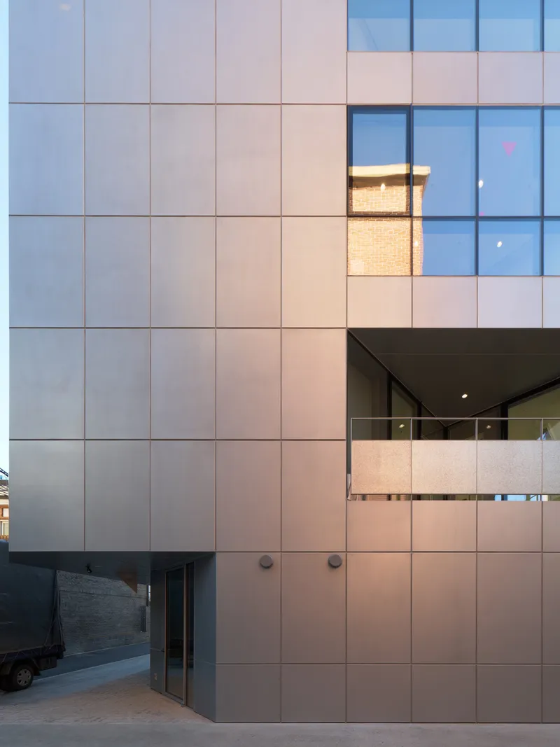 Facade grid of metal panels with recessed balcony and illuminated interior at dusk