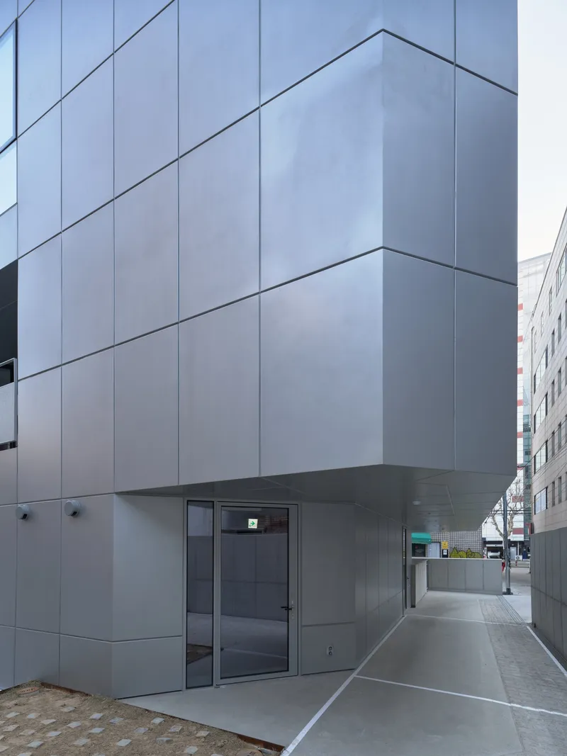 Exterior corner with pale grey metal panel facade, recessed glass entrance and covered walkway along the sidewalk