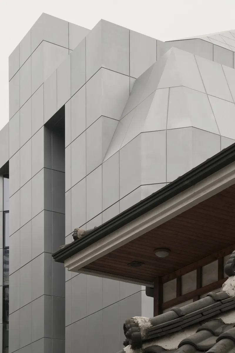 Faceted white panel cladding rising beside a tiled traditional roof under cloudy skies