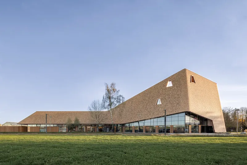 Side view of the shingled facade with triangular openings and glazed base across a lawn at dusk