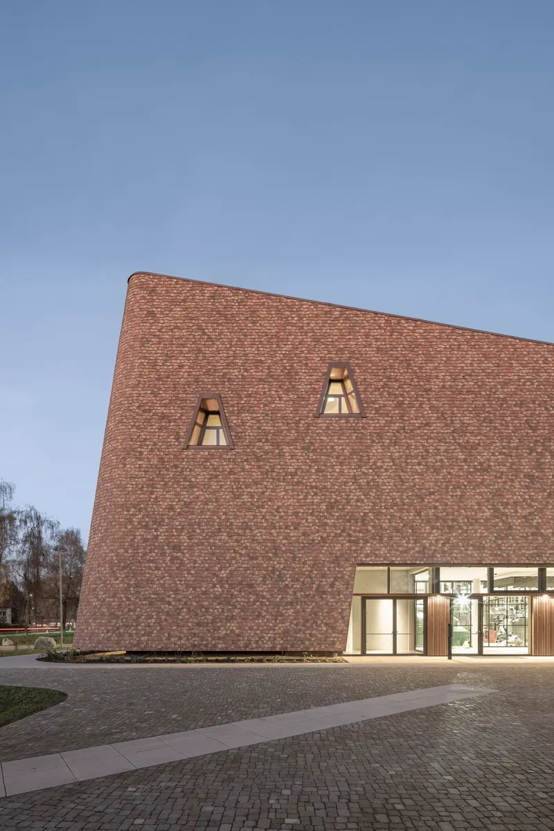 Brick facade with illuminated trapezoidal windows above the glazed entrance at twilight