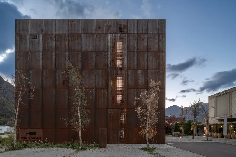 Weathered copper elevation with vertical perforated openings glowing at dusk beneath scattered clouds