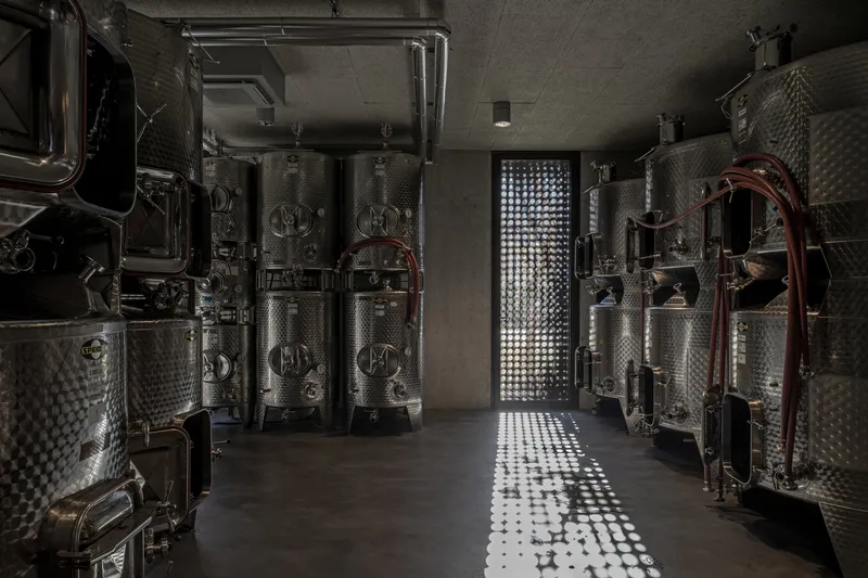 Stainless steel fermentation tanks flanking a corridor with dappled light from a perforated wall