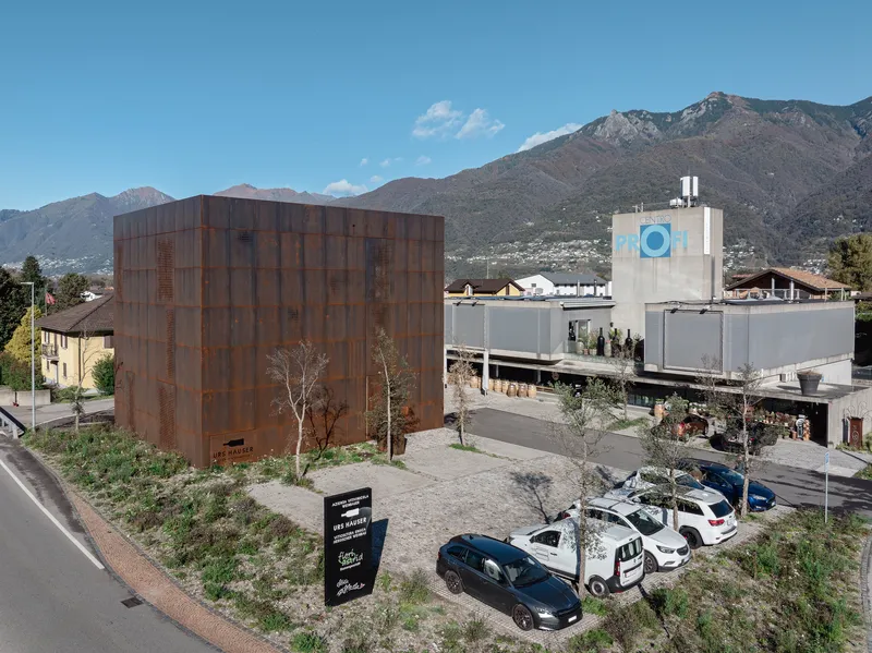 Weathered steel paneled cube adjacent to industrial buildings with mountains in the background
