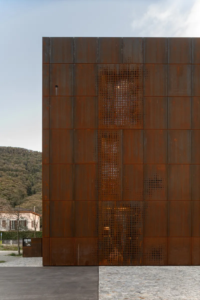 Weathered steel facade with vertical perforated panel against a hillside backdrop at dusk