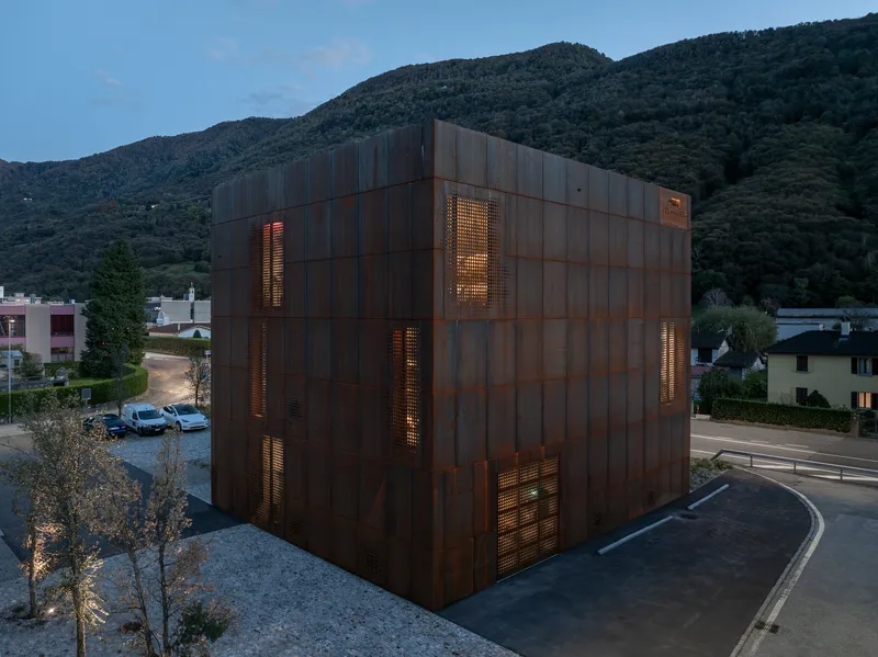 Corten steel cube with illuminated perforated openings at dusk against forested hillside backdrop