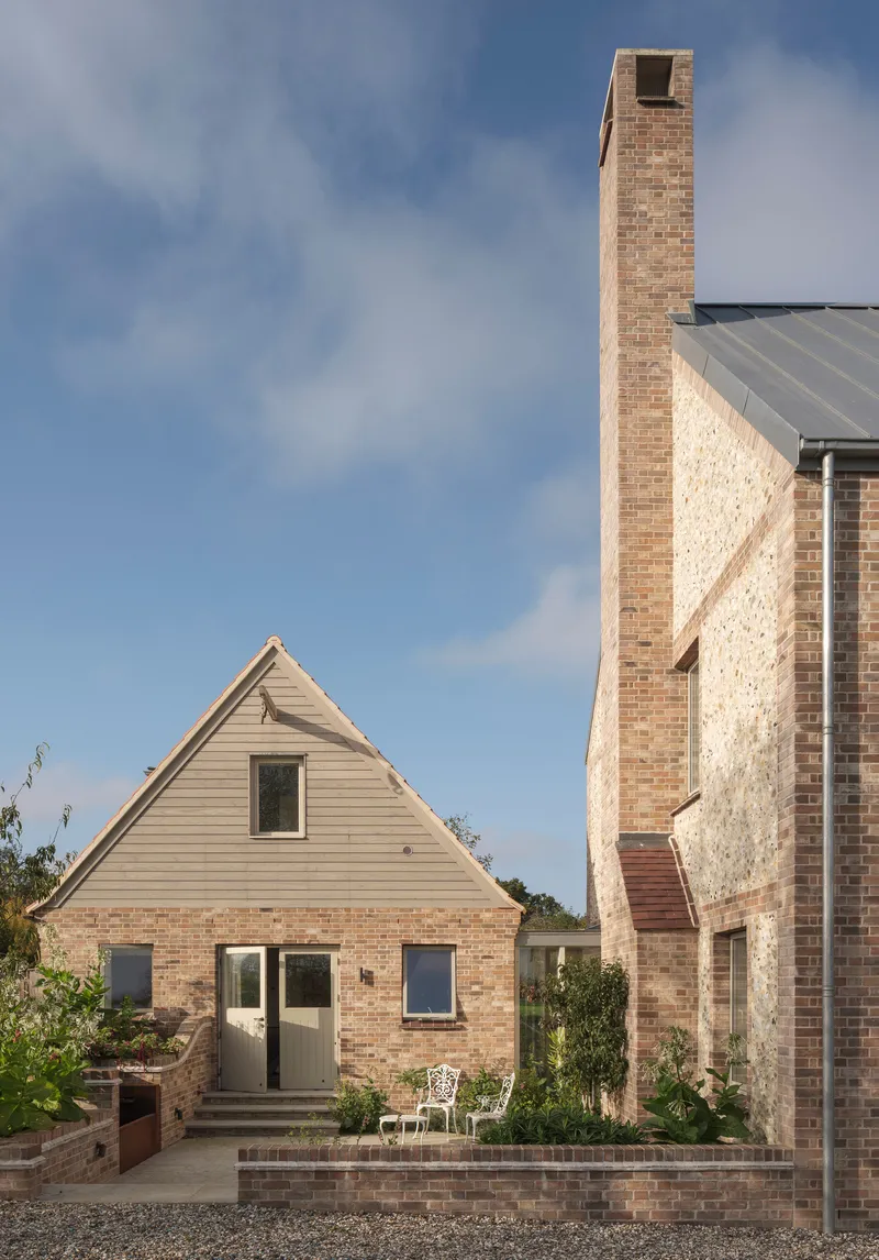 Brick-paved terrace between a pale timber-clad gable and tall stone chimney with planted beds
