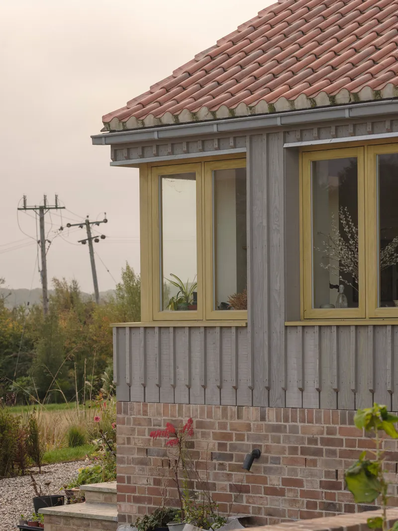 Corner detail of vertical timber cladding over brick base with terracotta tile roof and power lines in distance