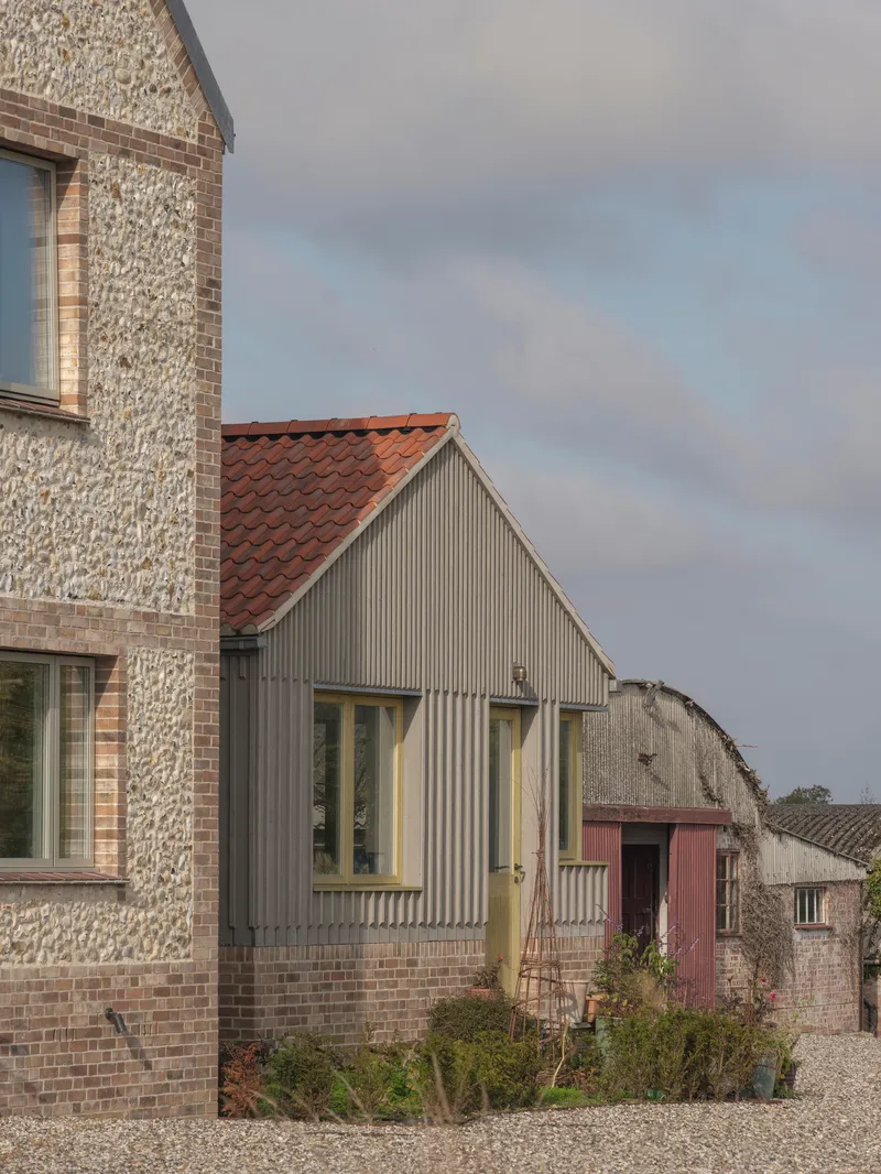 Gable end with vertical timber cladding and terracotta roof beside flint-faced volume and neighboring farm structures