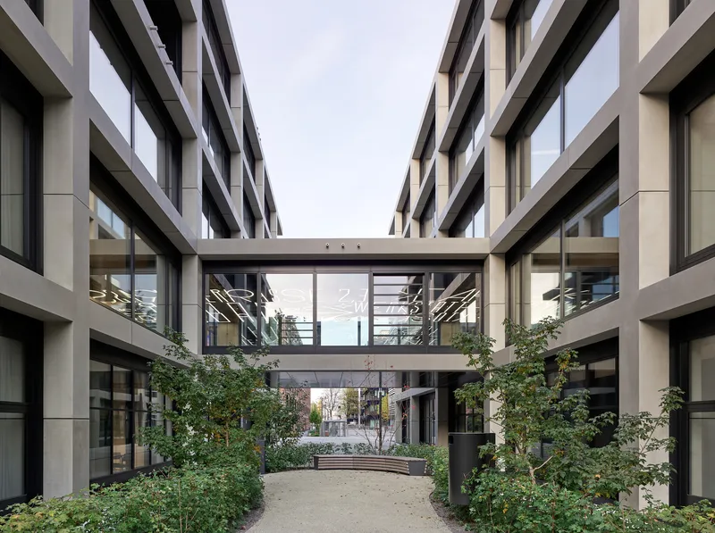 Landscaped courtyard with planted beds and trees beneath two cantilevered glass bridges connecting the wings