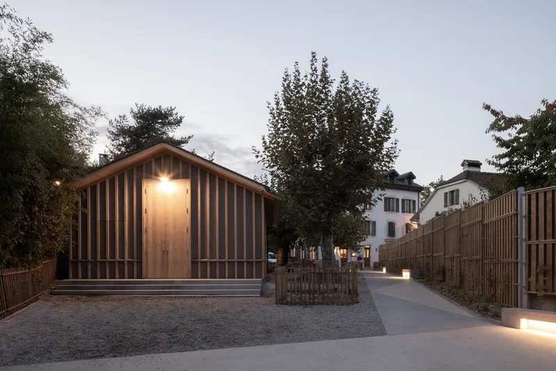Entry facade with vertical timber cladding illuminated at dusk beside a low wooden fence