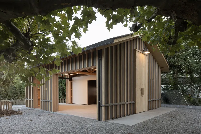 Low pavilion with alternating grey and timber vertical cladding beneath mature trees in autumn