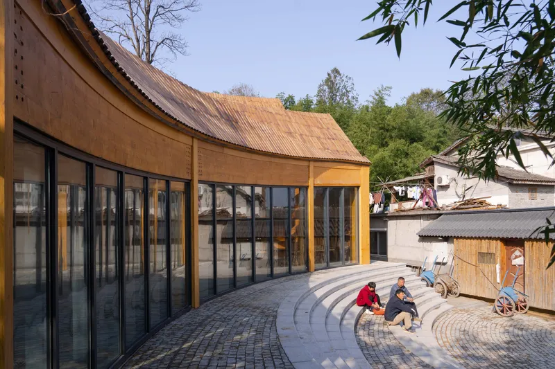 Curved corrugated metal roof over glazed facade and paved terrace with two seated visitors