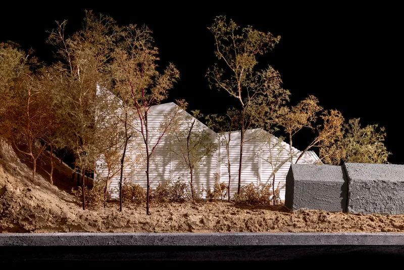 Side view of physical model showing illuminated ribbed facade behind bare trees and textured retaining wall