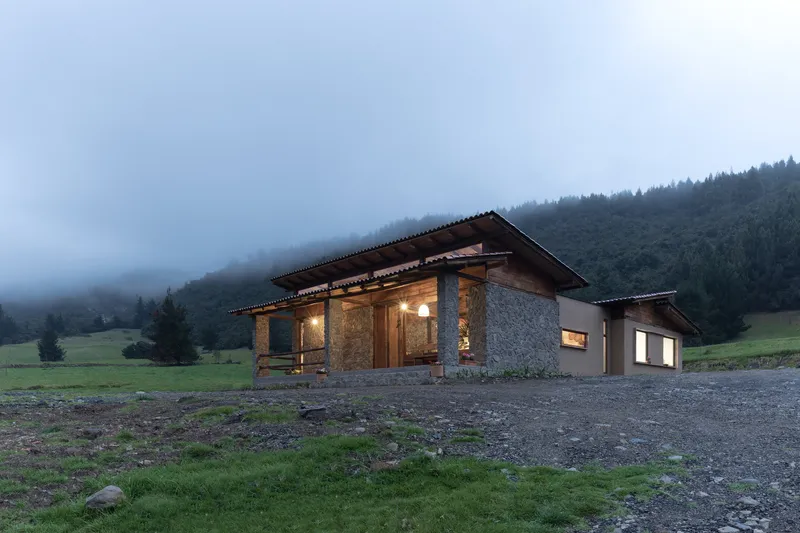 Stone and timber pavilion with illuminated interior recessed beneath wide roof at dusk in misty landscape