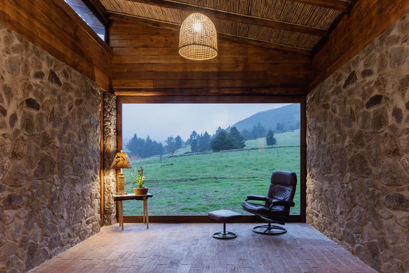 Stone walled room with woven bamboo ceiling and wicker pendant light framing view to misty pasture at dusk