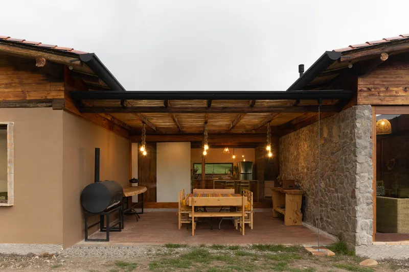 Covered courtyard with exposed timber beams and pendant bulbs connecting stone column and plastered walls at dusk