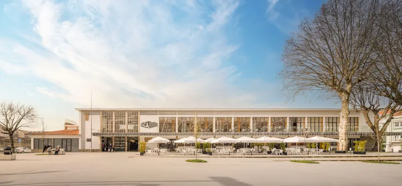 Frontal elevation of the glass-walled market hall facade with bare trees framing the entrance plaza