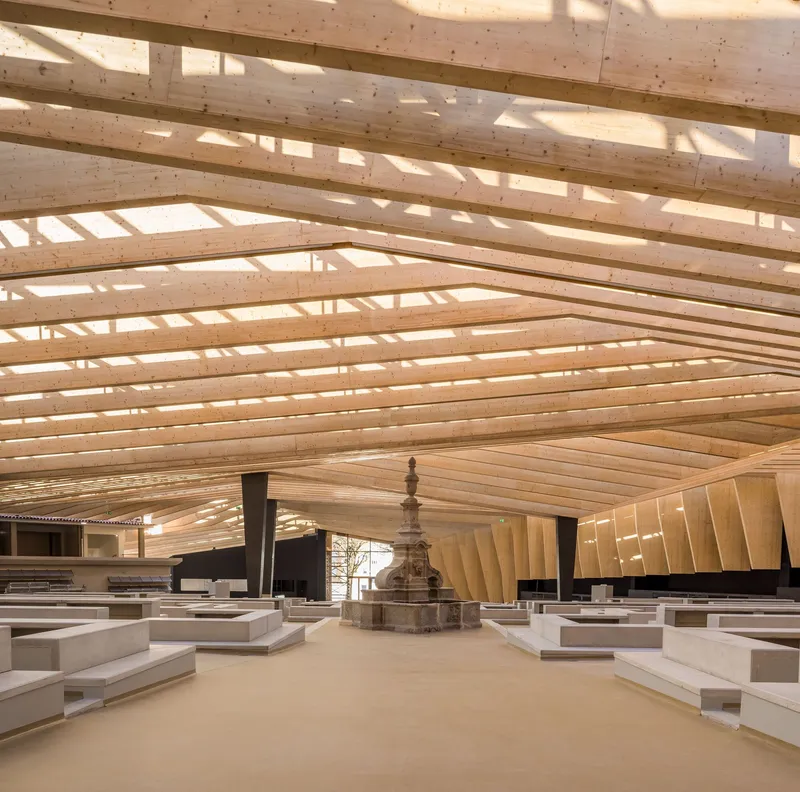 Central stone monument surrounded by stepped concrete seating under a layered timber roof with skylights