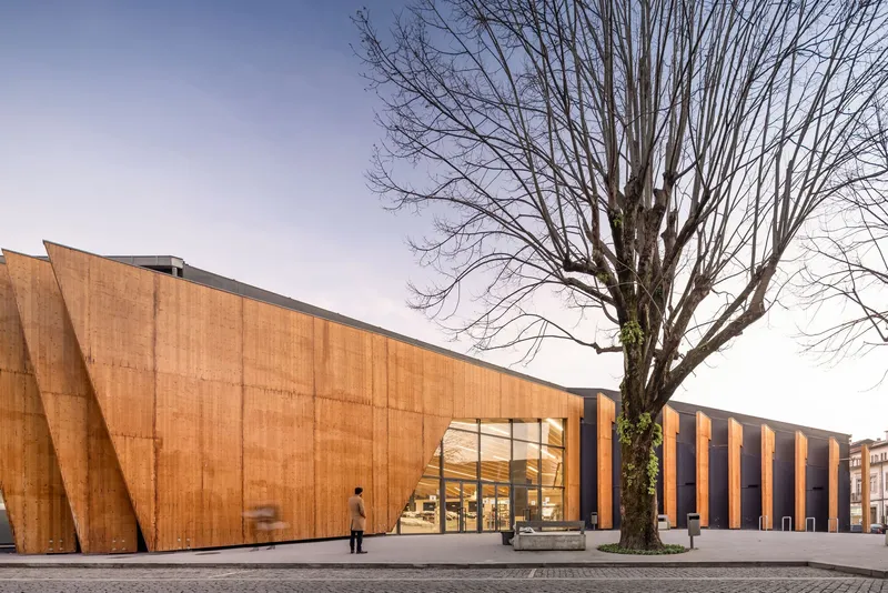 Angled timber facade with vertical fins beside a glazed entrance and bare winter tree at dusk