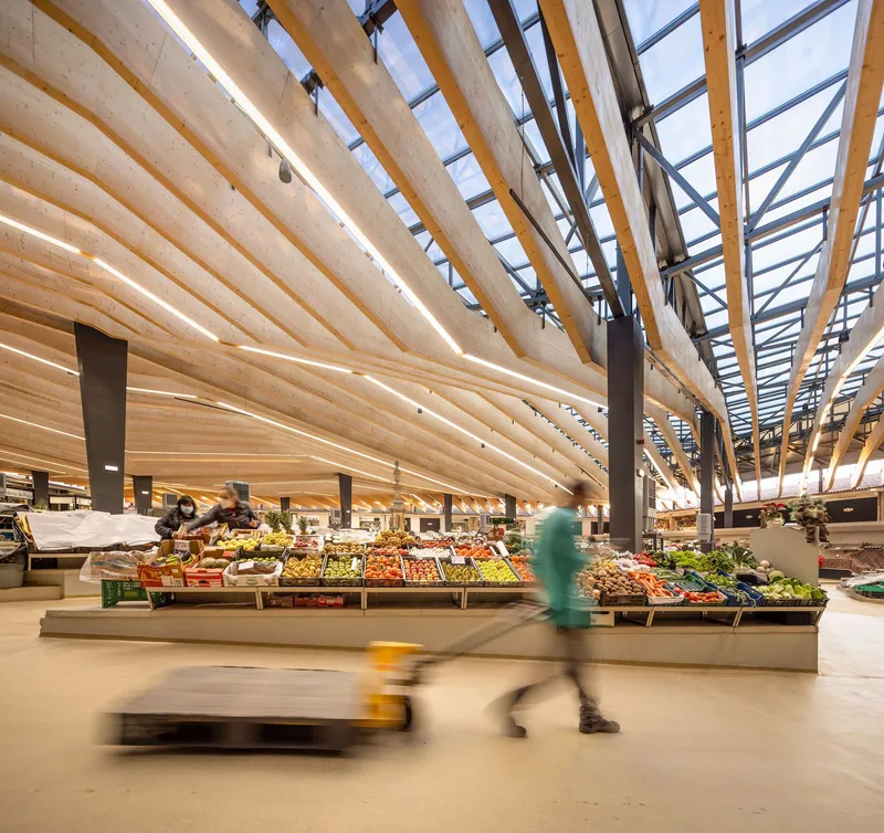 Market aisle with produce displays, curved timber ceiling, and shoppers in motion beneath skylights