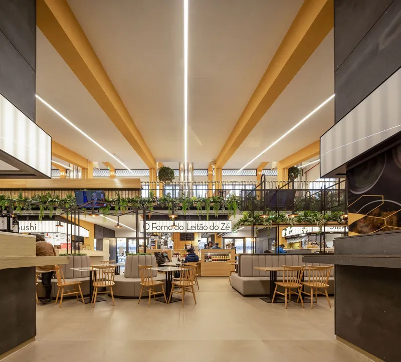 Double-height food hall interior with yellow laminated beams, mezzanine planters and linear recessed lighting