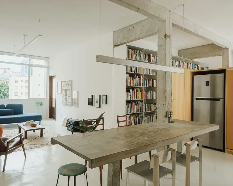 Open living and dining area with exposed concrete frame, bookshelf wall, and light from a large window