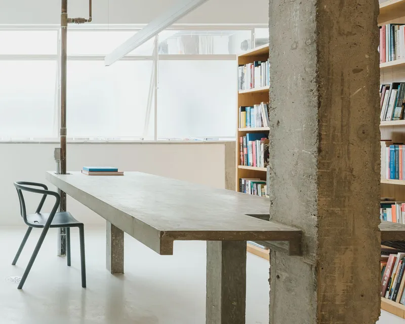 Concrete table on angled legs beside an exposed structural column and plywood bookshelf