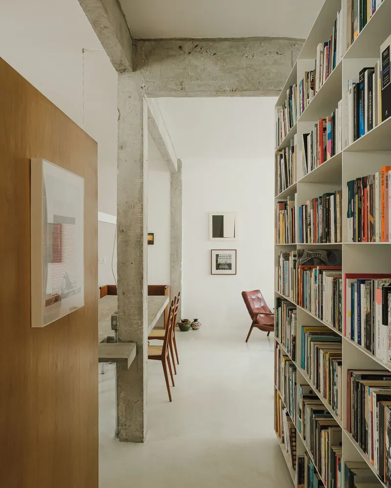 Corridor view past concrete columns toward white bookshelves and a red leather chair