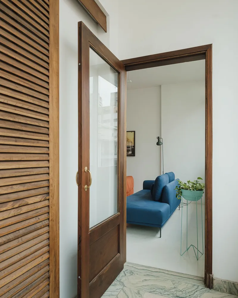 Dark timber-framed glass door opening from slatted screen hallway to living room with blue sofa