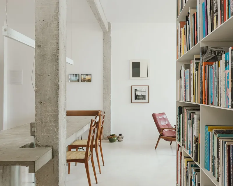 Double-height interior with exposed concrete columns, white walls, tall bookshelves and wooden dining chairs
