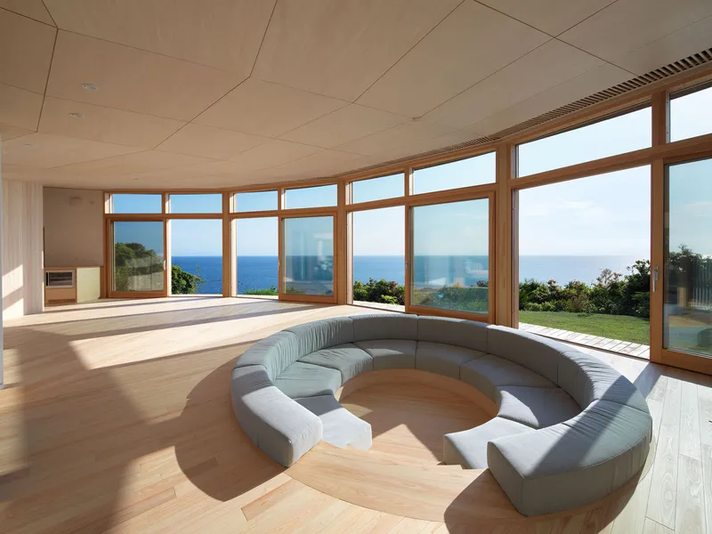 Curved living space with continuous timber-framed windows overlooking the ocean under a plywood ceiling