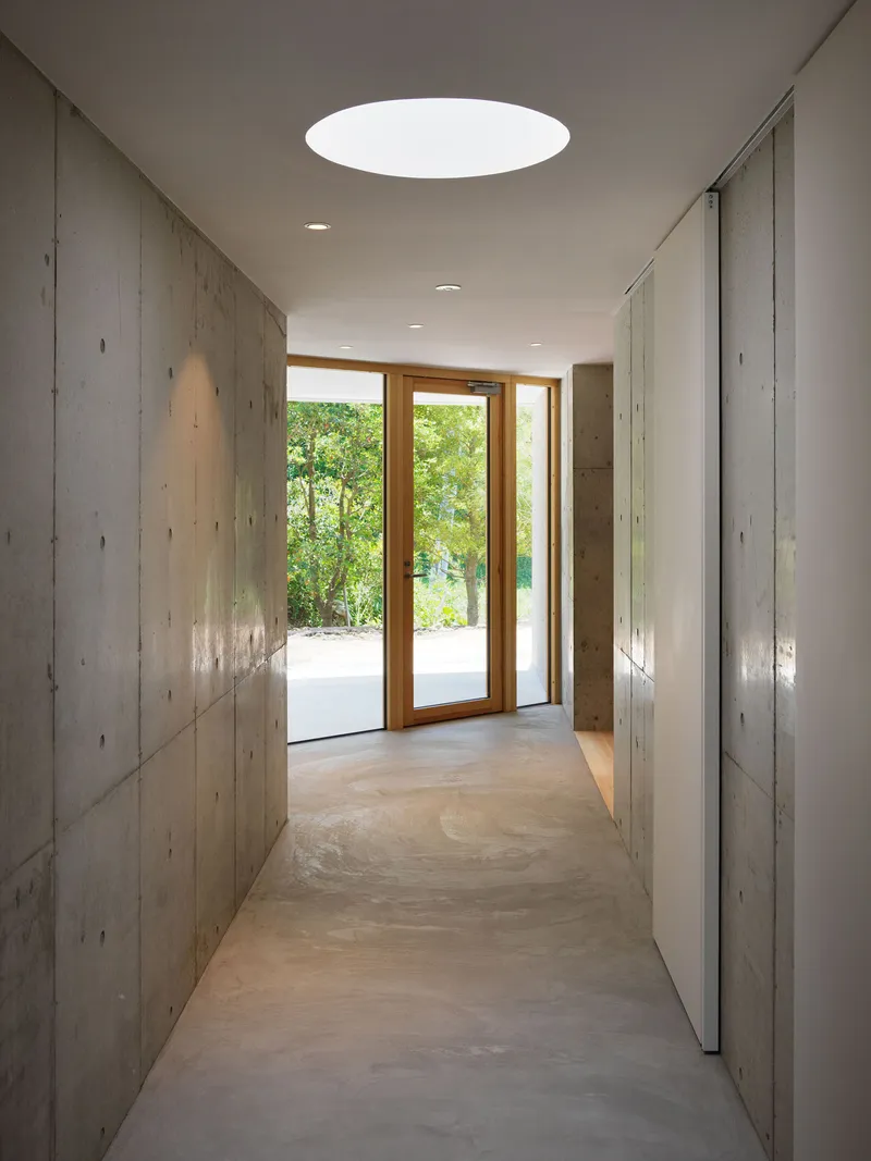 Concrete corridor with circular skylight and timber-framed doors leading to garden