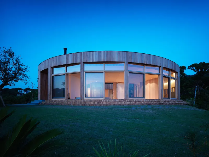 Curved timber facade with full-height glazing across lawn under clear blue twilight sky