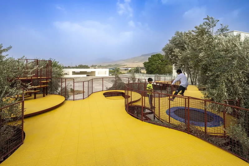 Curved yellow platform with children playing among red mesh barriers and surrounding olive trees